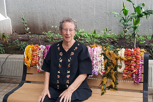Photograph of Jan Dapitan sitting on a bench dedicated in her behalf for the role she played in establishing Hawaii's Environmental Court. The top of the bench is covered with leis.