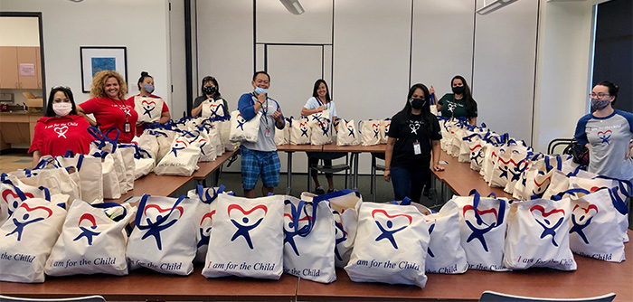 Nine CASA staff members in a large room stand beside tables stacked with CASA care bags, May 2020.
