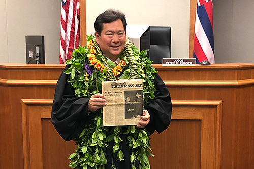 Third Circuit Court Judge Peter K. Kubota stands in front of his courtroom bench holding a copy of the 04/17/1974 edition of the Hawaii Tribune-Herald newspaper.