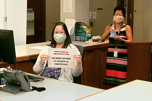 Librarian Marlene Cuenco and State Law Librarian Jenny R.F.F. Silbiger wearing masks and holding up hand sanitizer and the reservations phone number at the Hawaii Supreme Court Law Library reference desk, 08/03/2020.