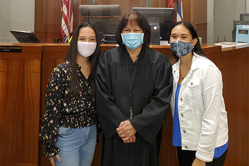 A woman judge wearing a black robe and a face mask posing with two young women in front of the judges bench
