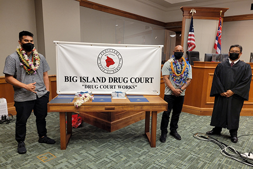 In the courtroom, two graduates stand on the left and right sides of a table holding a Big Island Drug Court banner (“Drug Court Works”), with Hilo Drug Court Judge Henry Nakamoto on the far right, at the program’s 57th graduation ceremony, 08/05/2021.