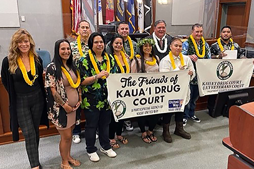 Kauai Drug Court graduates and Deputy Chief Judge Michael K. Soong pose with Friends of Kauai Drug Court and Veterans Court signs in front of the courtroom bench, 05-23-2022.