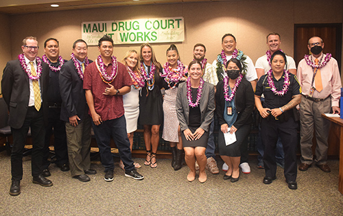 Maui Drug Court team members and seven graduates stand together in the courtroom in front of a sign that says, "Maui Drug Court Works," at the 05/19/2022 graduation ceremony.
