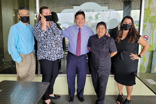 Deputy Prosecutor Kevin Hashizaki, Drug Court Supervisor Pagan Devela, Judge Peter Kubota, Short and Sweet Owner Maria Short, and Deputy Public Defender Sheri Tavares stop for a picture together in front of the window of Short and Sweet, 05/26/2022.