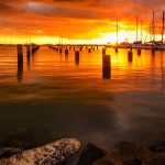 Photograph looking out over the water, with sailboats docked, sails down, orange and gray on the water and clouds above, bright yellow in the center where the sun is setting.