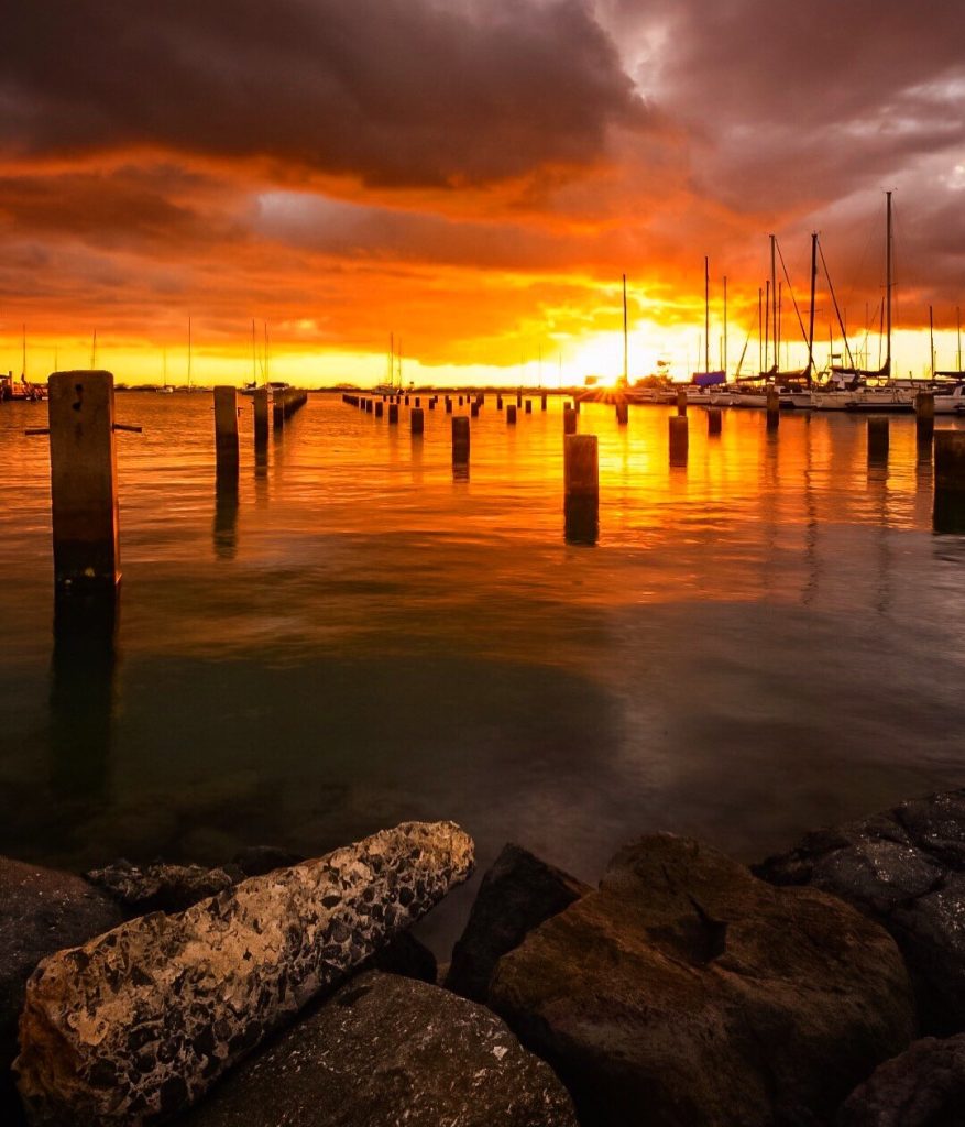 Photograph looking out over the water, with sailboats docked, sails down, orange and gray on the water and clouds above, bright yellow in the center where the sun is setting.