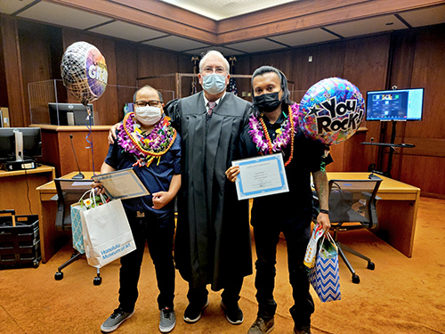 Two DWI Court graduates adorned with lei, wearing masks to protect against COVID-19, pose with DWI Court Judge Alvin Nishimura in the Honolulu District Court courtroom, September 8, 2022.