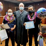 Two DWI Court graduates adorned with lei, wearing masks to protect against COVID-19, pose with DWI Court Judge Alvin Nishimura in the Honolulu District Court courtroom, September 8, 2022.