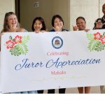 Six smiling Judiciary employees stand outside the Circuit Court building on Punchbowl Street in Honolulu holding a banner that reads celebrating juror appreciation.