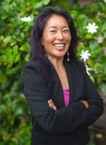 The Hawaiʻi State Judiciary’s State Law Librarian and Access to Justice Coordinator, Jenny Silbiger, stands for a photo in front of a hedge with green leaves and white flowers, 04-26-2025.