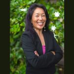 The Hawaiʻi State Judiciary’s State Law Librarian and Access to Justice Coordinator, Jenny Silbiger, stands for a photo in front of a hedge with green leaves and white flowers, 04-26-2025.