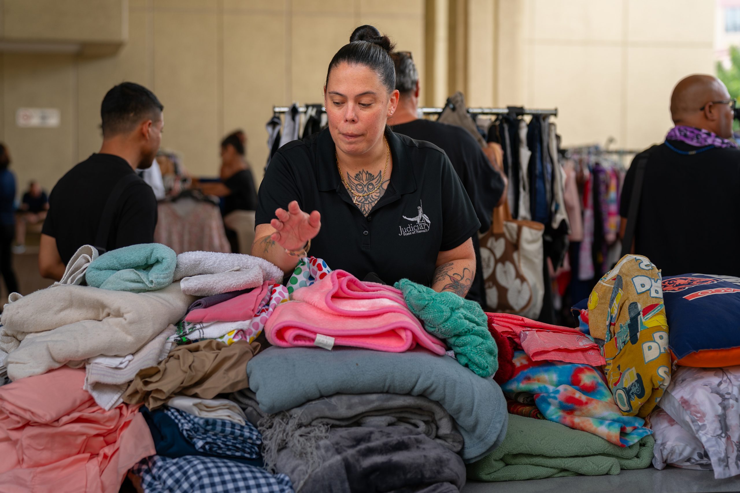 Senior Probation Officer Jamie Logan arranges stacks of folded clothing and blankets at a donations table during a service fair, with other attendees and booths visible in the background.