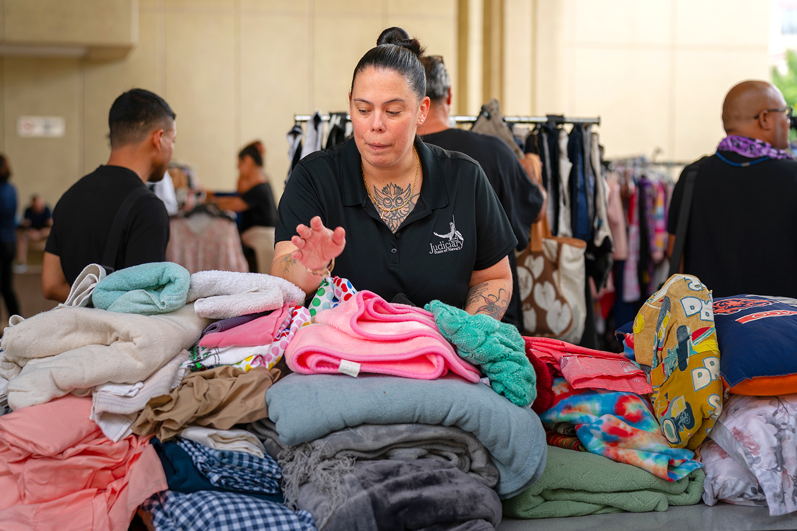 Senior Probation Officer Jamie Logan arranges stacks of folded clothing and blankets at a donations table during a service fair, with other attendees and booths visible in the background.